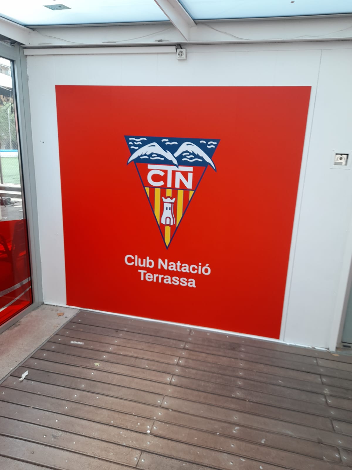 Mural corporativo Club Natació Terrassa con escudo CTN sobre fondo rojo instalado en acceso exterior