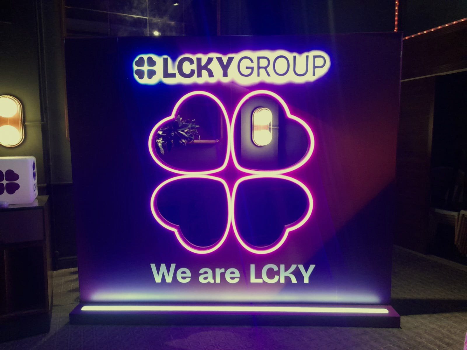Backdrop corporativo LCKY Group con trébol de neón rosa magenta y panel azul oscuro con claim We are LCKY